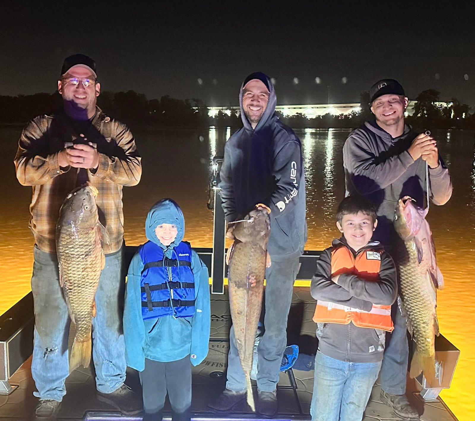 Group of anglers with kids holding carp and catfish on the boat at night